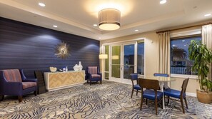 Ein modernes Hotelzimmer mit einem gemusterten Teppich, einem runden Tisch mit Stühlen, einer Couch, einem Schrank und einem großen Fenster mit Vorhängen.