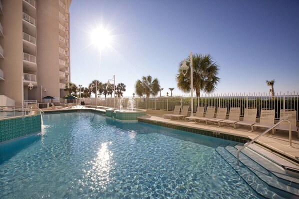 A swimming pool with a sunlit sky, a white fence, and palm trees.