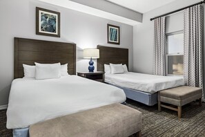A hotel room with two beds, a bench, a nightstand with a lamp, and a window with patterned curtains.