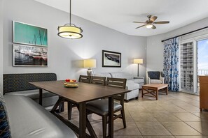 A modern living room with a dining table, sofa, and chairs, a ceiling fan, and large windows with curtains.