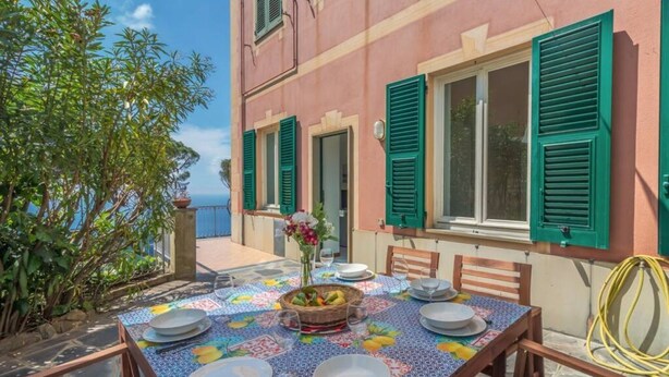 A dining table set outdoors with a view of the sea, a building with green shutters, and a garden area. - CASA FLAMINIA - Panorama, Relaxation, and Parking (Ruta)