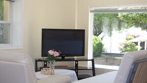 Une pièce comprenant un lit, un téléviseur posé sur un meuble, une petite table avec un vase de fleurs et une fenêtre donnant sur un paysage verdoyant.