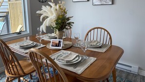 Ein Esszimmer mit einem Holztisch, gedeckt für vier Personen, umgeben von Holzstühlen. Auf dem Tisch steht eine Vase mit weißen Blumen, und an der Wand hängen zwei gerahmte Kunstwerke.