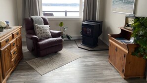 Ein gemütliches Wohnzimmer mit Ledersessel, Holzkommode, Kaminofen und großem Fenster mit Blick auf das Wasser.