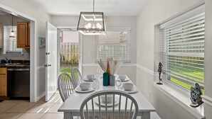 A dining area with a white table set for four, a hanging light fixture, and a window with blinds. - Three Minutes to White Sand Beach! (Panama City Beach)
