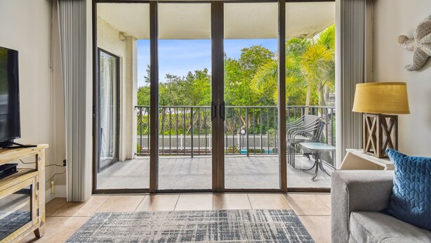 A living room with a television, a sofa, a lamp, and a view of a balcony with outdoor furniture. - Waterfront views - steps from the Beach! (Indian Shores)