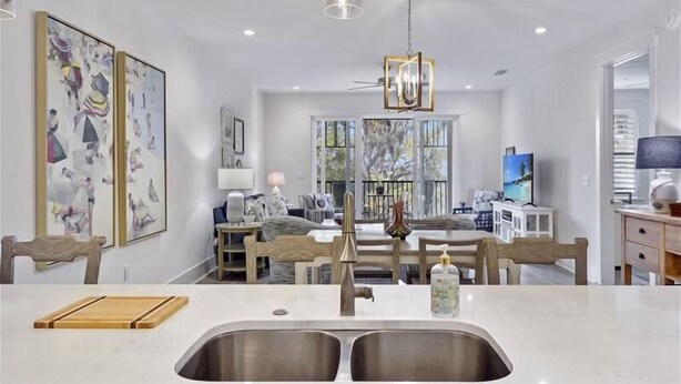 A modern kitchen with a double sink, wooden chairs, and a dining table. There are framed artworks on the wall and a television in the background. - The Moorings #22, Your perfect getaway starts here! (Jekyll Island)