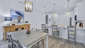 A modern kitchen with white cabinetry, a dining area with a wooden table and chairs, pendant lighting, and a large framed picture on the wall. - The Moorings #22, Your perfect getaway starts here! (Jekyll Island)