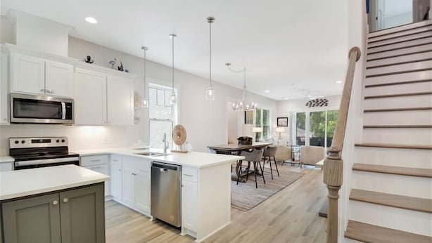 A modern kitchen with white cabinetry, a built-in oven, and a dishwasher. There is a dining area with a table and chairs, and a staircase with wooden steps. - The Moorings 43 Relax in Luxury - Pet Friendly (Jekyll Island)