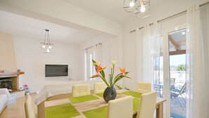 A modern dining area with a wooden table, green placemats, and white chairs. There is a vase with flowers on the table, and a flat-screen TV mounted on the wall. - Villa Telio: Sunny pool, A/C, WiFi, BBQ (Sgompou)