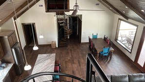 A spacious loft with a high ceiling, a mezzanine level, a dining area with a wooden table and chairs, and a large window providing a view of the outdoors. - The River Lodge (Uvalde)