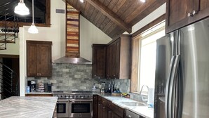 A modern kitchen with dark wooden cabinets, a stainless steel refrigerator, a large island with a marble countertop, and a built-in oven. - The River Lodge (Uvalde)