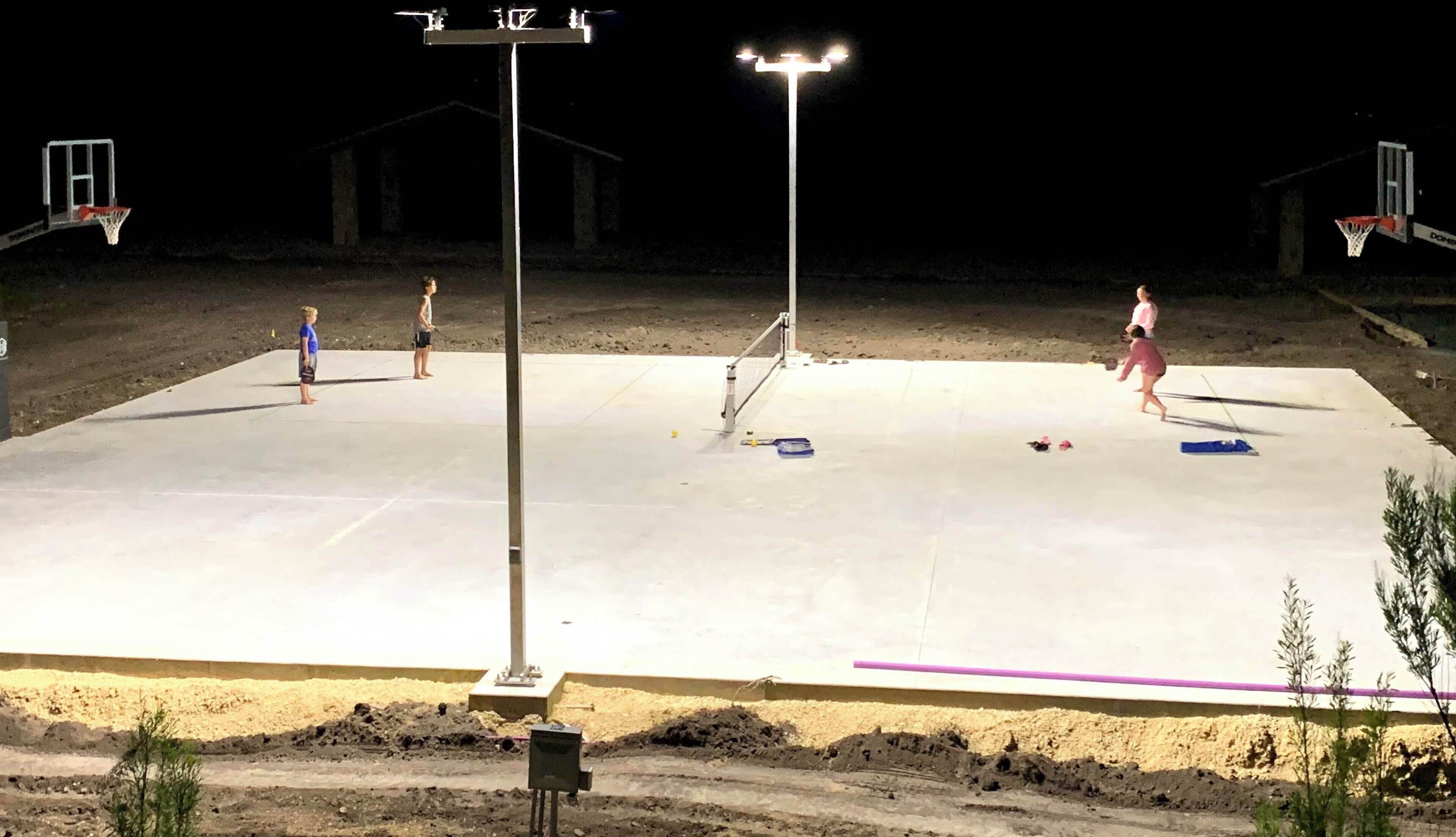 An outdoor basketball court at night with four people playing.