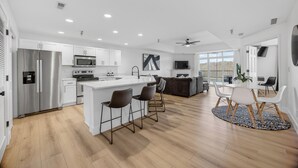 A modern kitchen with a central island, bar stools, a dining area with a round table and chairs, and a large window offering a view.