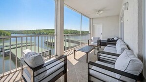 A balcony with a view of a lake and greenery, furnished with a sofa and chairs.