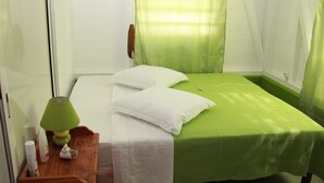 Une petite chambre avec un lit simple, une couvre-lit verte, des oreillers blancs, une table de chevet avec une lampe verte et une petite commode où sont rangés les articles de toilette.