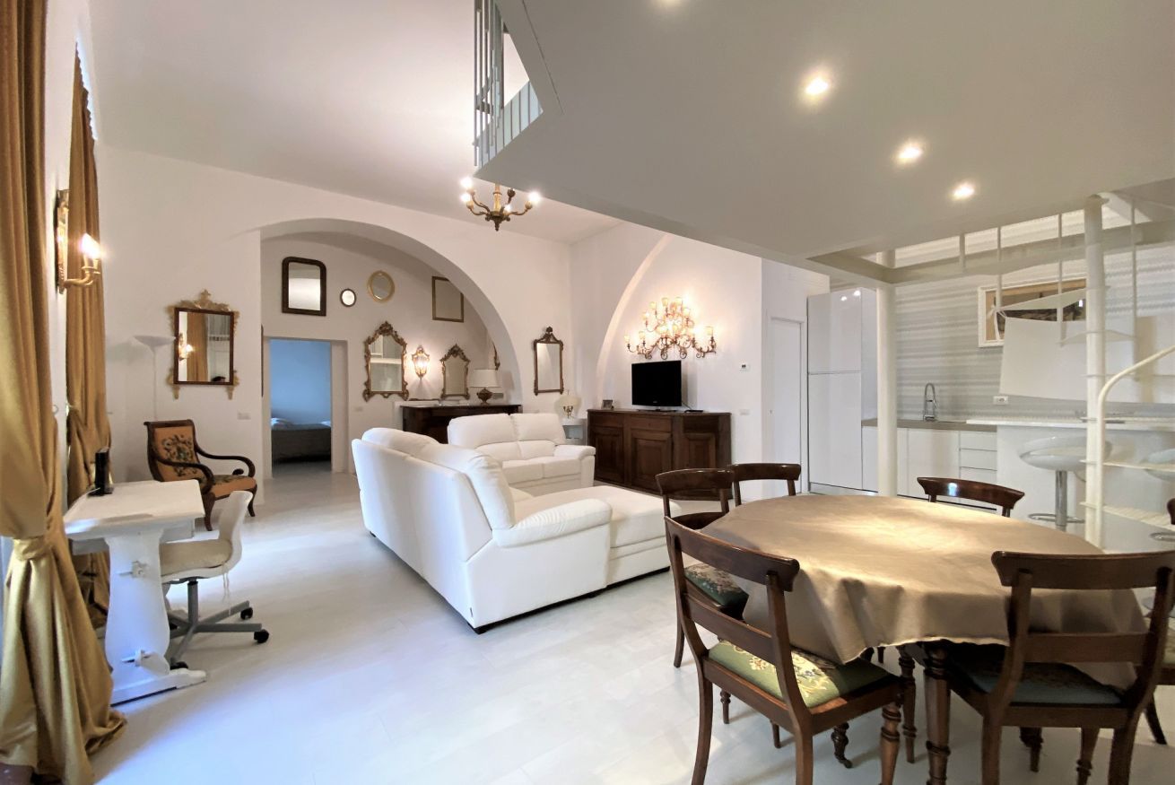 A spacious living area with a white sofa, a dining table with chairs, and a staircase leading to an upper level.
