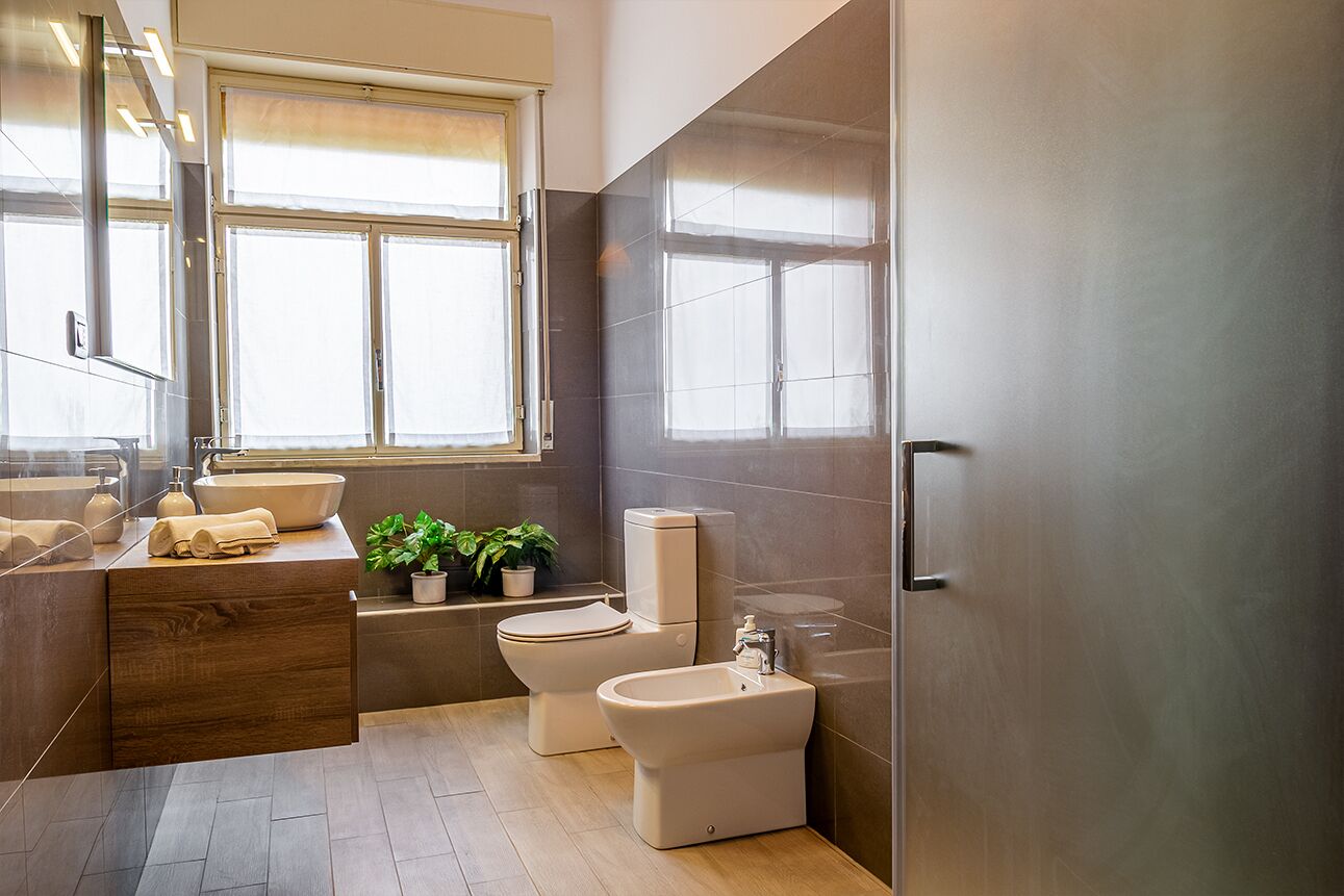 Une salle de bain moderne avec un meuble-lavabo en bois, un évier, des toilettes, un bidet et une grande fenêtre qui laisse entrer la lumière naturelle.