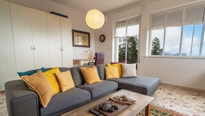 A modern living room with a grey sectional sofa, a coffee table with decorative stones and a remote control, a patterned rug, and a large window with blinds. - Villa Alberto - Country Relax (Puntalazzo)