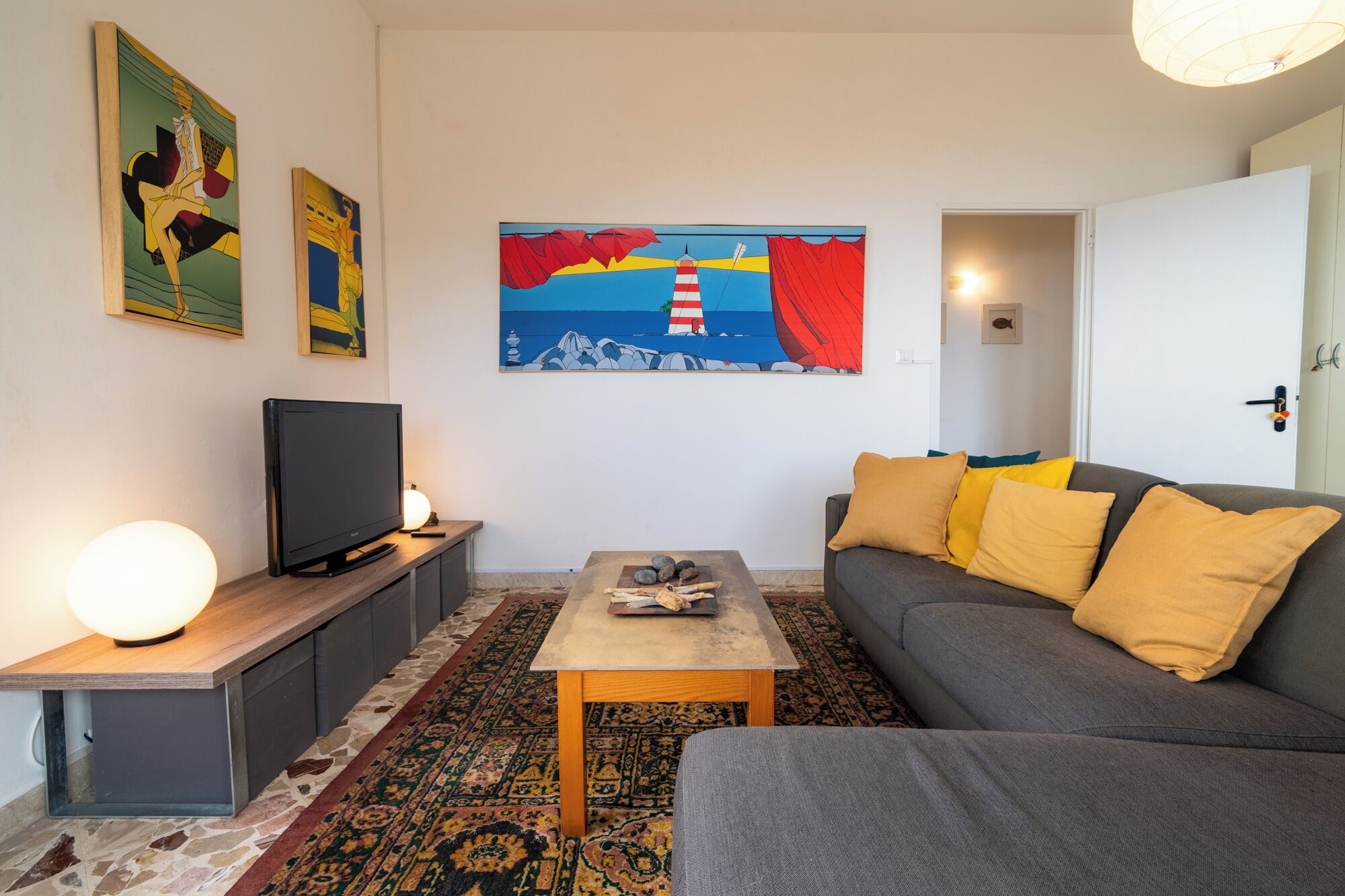 A modern living room with a grey sofa, a wooden coffee table, and a flat-screen TV on a wooden stand. There are colorful paintings on the wall and a patterned rug on the floor.
