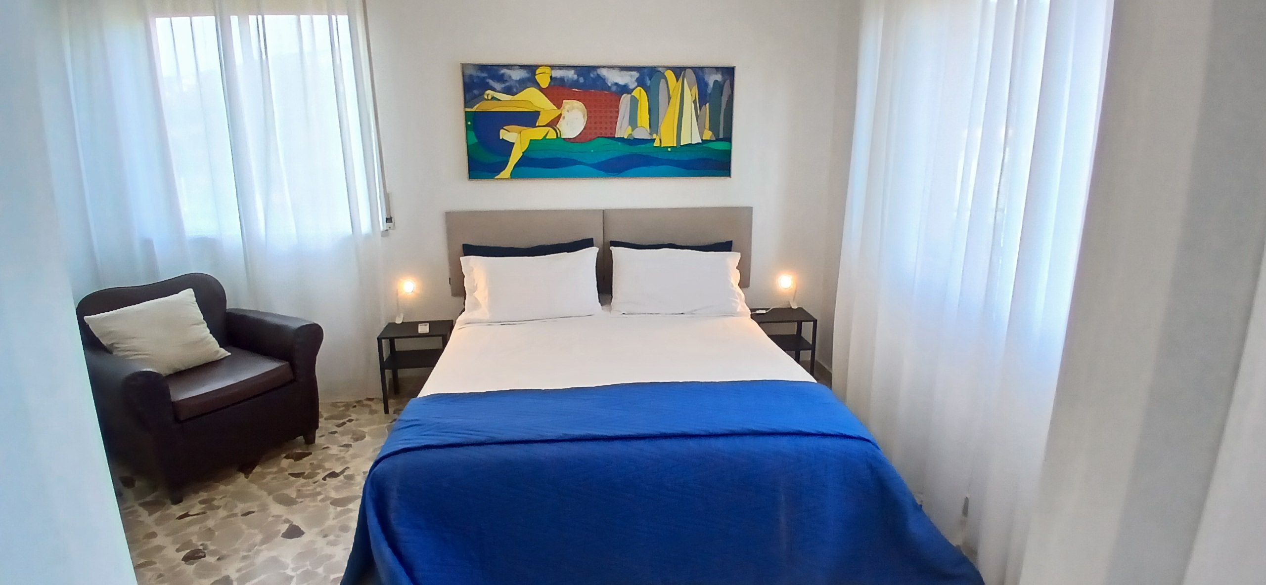 A hotel room with a bed, a chair, a nightstand, and a painting on the wall.