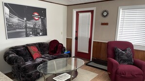 Un salon avec une table basse en verre, un fauteuil rouge, un canapé noir, une affiche encadrée provenant du métro et une porte rouge.