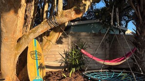 A hammock hanging from a tree, a surfboard leaning against a tree, and a wooden fence in the background.