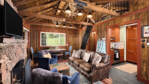 A rustic cabin interior with a stone fireplace, leather sofa, blue chairs, a dining area, and a kitchen.