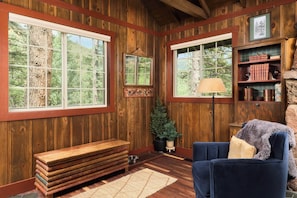 Interior acogedor de una cabaña con paredes de madera, ventanas amplias, un sofá azul, un banco y un pequeño árbol.