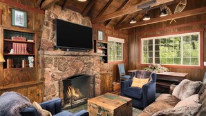 A cozy cabin interior with a stone fireplace, wooden walls, and a large window offering a view of the outdoors.
