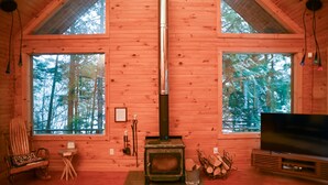 Ein gemütliches Blockhaus-Interieur mit Kaminofen, großen Fenstern, Fernseher und Schaukelstuhl.