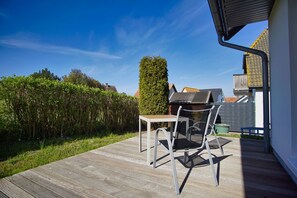 A wooden deck with a table and chairs, surrounded by a hedge and a tall tree. - Die Ferienwohnung Dünenrose im Haus Südperd - Haus Südperd (Mönchgut)