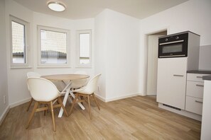 A compact kitchen with a dining area, featuring a round wooden table and white chairs, a built-in oven, and a window with a view of a building. - Die Ferienwohnung Dünenrose im Haus Südperd - Haus Südperd (Mönchgut)