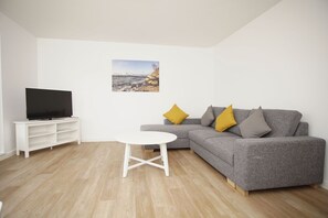 A living room with a grey sofa, a white coffee table, a flat-screen TV on a white stand, and a painting of a beach scene. - Die Ferienwohnung Dünenrose im Haus Südperd - Haus Südperd (Mönchgut)
