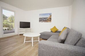 A modern living room with a grey sofa, white coffee table, and a flat-screen TV on a white stand. - Die Ferienwohnung Dünenrose im Haus Südperd - Haus Südperd (Mönchgut)