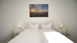 A neatly made bed with white linens and two bedside tables with lamps, set against a plain wall with a landscape painting above. - Haus Südperd Ferienwohnung Strandwinde - Haus Südperd (Mönchgut)