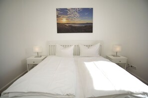 Una cama bien hecha con sábanas blancas y dos mesitas de noche con lámparas, frente a una pared lisa con un cuadro de paisaje.