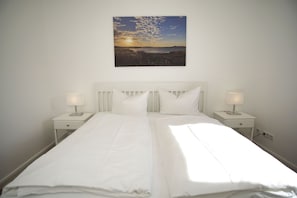 A neatly made bed with white linens and two bedside tables with lamps, set against a plain wall with a landscape painting above. - Haus Südperd Ferienwohnung Strandwinde - Haus Südperd (Mönchgut)