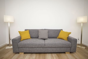 A grey sofa with two mustard yellow pillows and a grey pillow, flanked by two floor lamps with white shades, in a room with wooden flooring.
