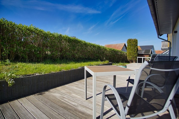 An outdoor deck with a table and chairs, surrounded by a hedge and a view of a house.