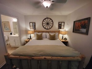 Une chambre à coucher avec une tête de lit en bois, un grand lit avec de nombreux oreillers, des lampes de chevet, un élément décoratif rond fixé au mur, un tableau encadré et une salle de bain visible à travers une porte ouverte.