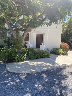 A white building with a dark door, surrounded by greenery and a gravel driveway.