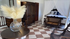 Una habitación con suelo a cuadros, una mesa redonda con un jarrón, un armario de madera, una cama con dosel y un sofá de mimbre.