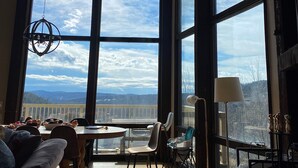 Il s’agit d’une pièce spacieuse, dotée de grandes fenêtres qui offrent une vue sur les montagnes. On y trouve une table à manger avec des chaises, un canapé et une lampe.