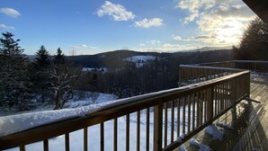 Une terrasse en bois, dont les rampes sont recouvertes de neige, offre une vue sur un paysage enneigé, avec des arbres et des collines lointaines.