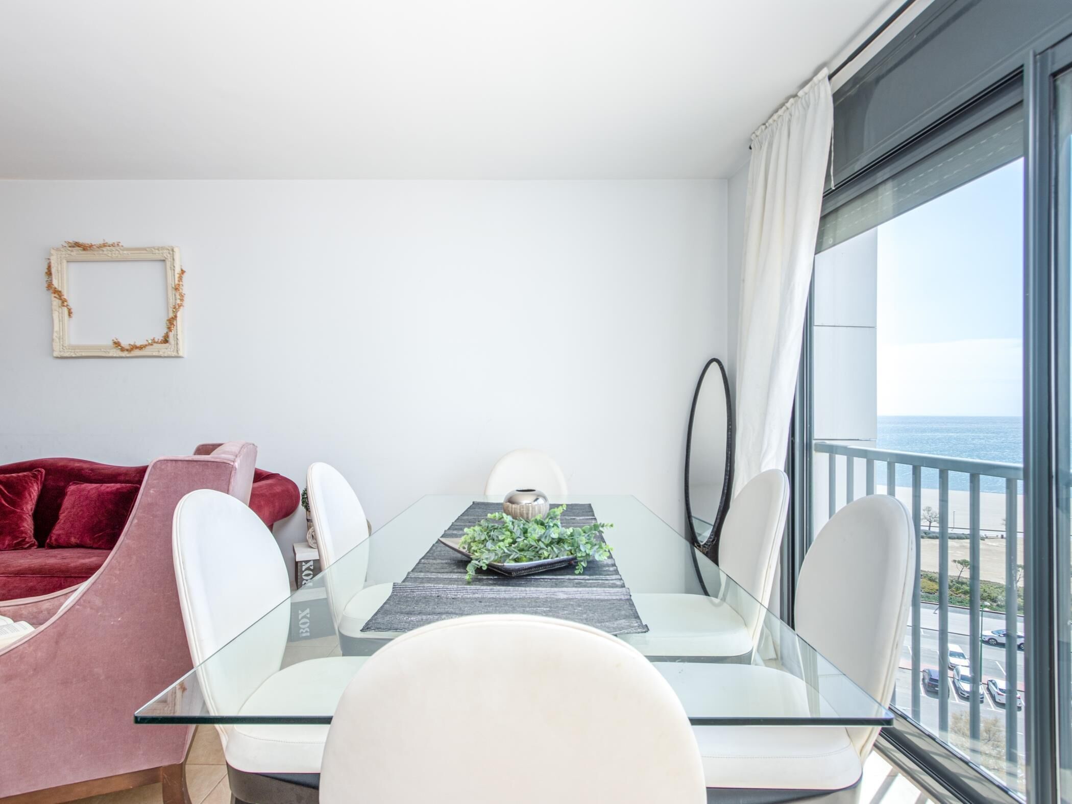 A modern dining area with a glass table, white chairs, and a view of the ocean through large windows.