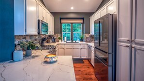 Cocina moderna con armarios blancos, encimera de mármol y una ventana grande con vistas a árboles.