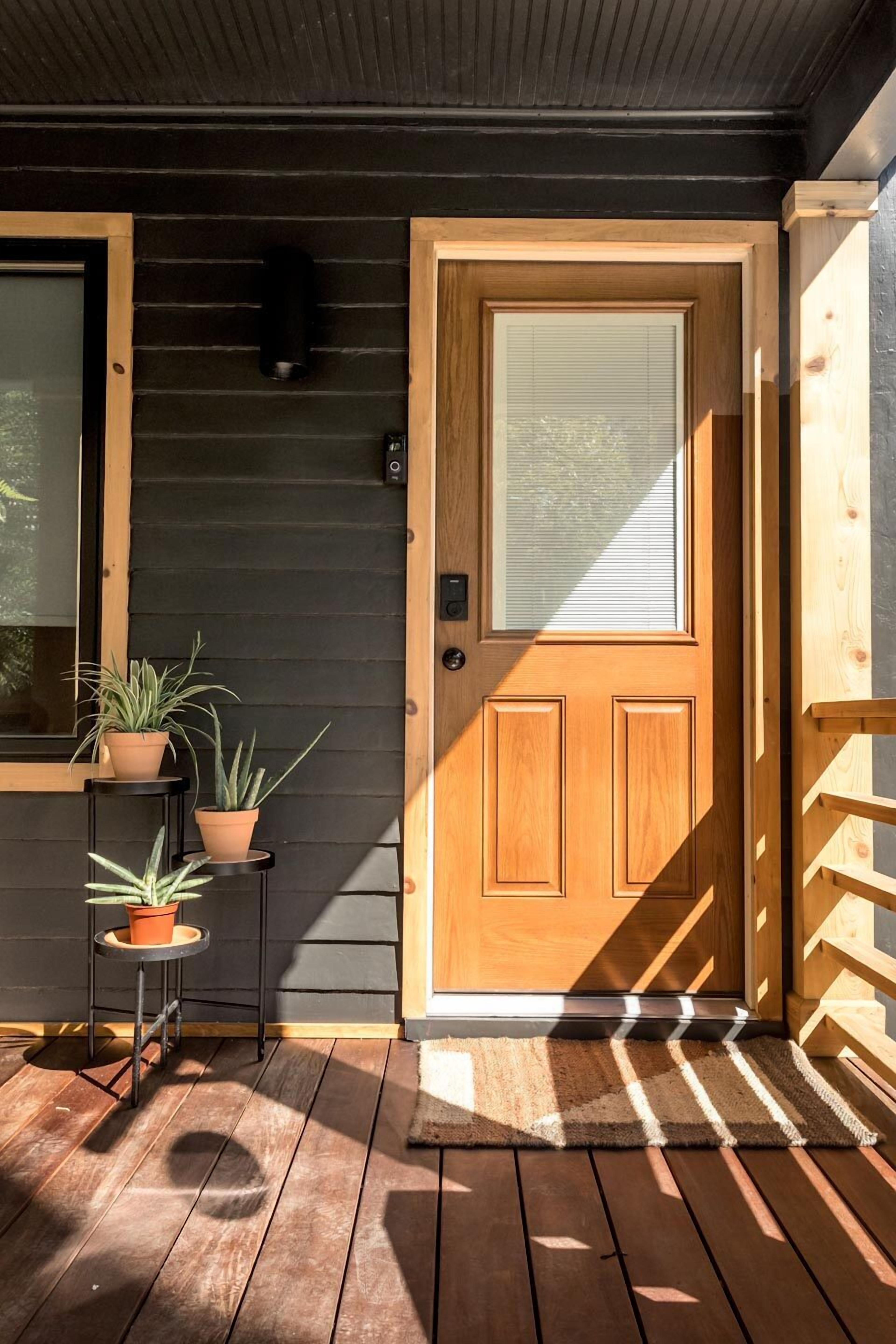 Une porte en bois avec un panneau de verre, une petite étagère extérieure où sont posées des plantes en pot, et une terrasse en bois.