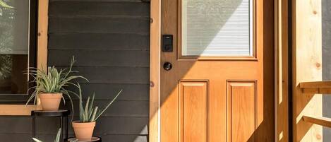 Une porte en bois avec un panneau de verre, une petite étagère extérieure où sont posées des plantes en pot, et une terrasse en bois.