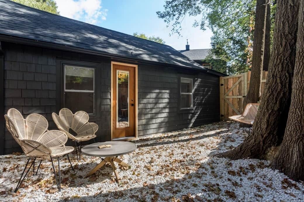 Una casa con fachada oscura, una puerta de madera y un patio con sillas de mimbre y una mesa.